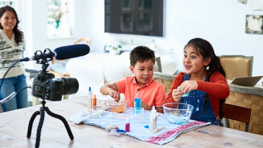 Two young siblings conduct a home science experiment in front of a camera at the dining table while their mum looks on