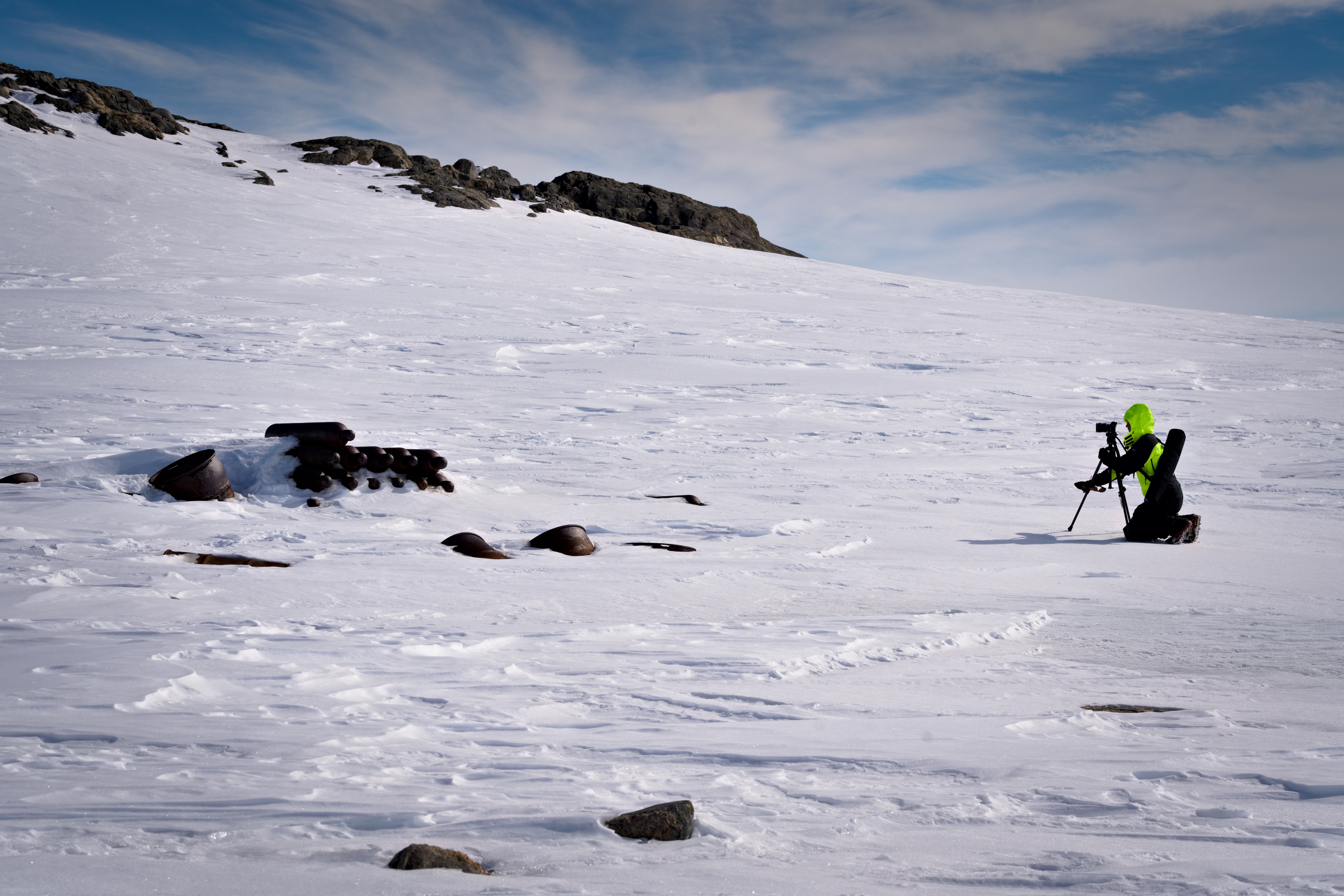Polly Stanton usa equipamento de neve preto e amarelo fluorescente e se ajoelha atrás de uma câmera em um tripé no gelo da Antártica.