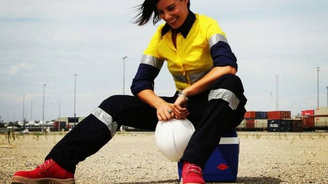 A woman wears a yellow and blue long sleeved work shirt, high-visibility cargo pants and red work boots.