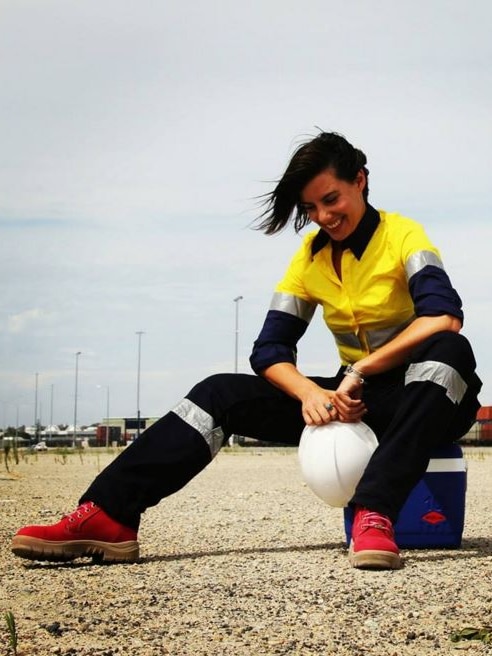 A woman wears a yellow and blue long sleeved work shirt, high-visibility cargo pants and red work boots.