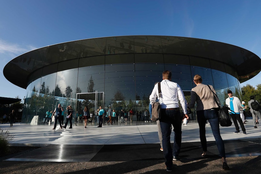 Smartly dressed people walk towards a modern, circular building on a sunny day.