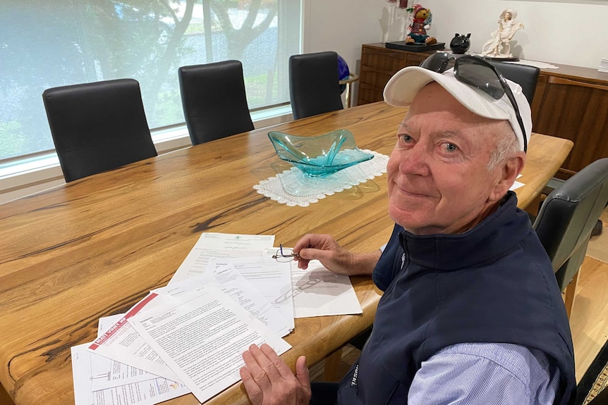 An elderly man wearing a hat sitting at a table with some documents looking over his shoulder towards the camera