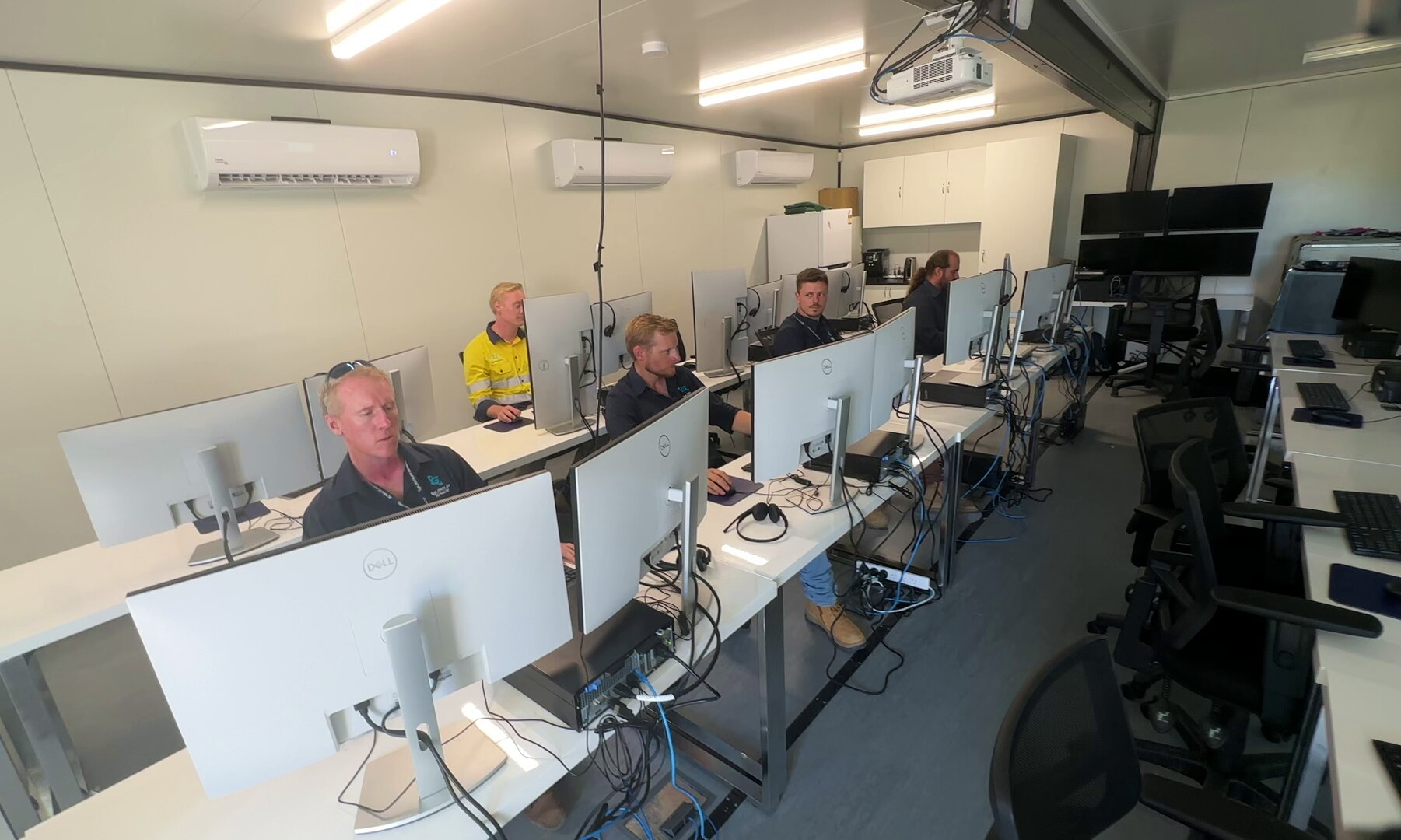 Four people sit behind computers at a long desk.
