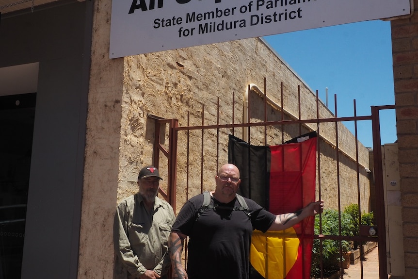 Darren Perry and Robby Wirramanda Knight stand next to a black, red and yellow Aboriginal flag hung on a gate.