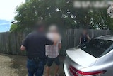 A police officer escorts a man from a property. Their faces are blurred.
