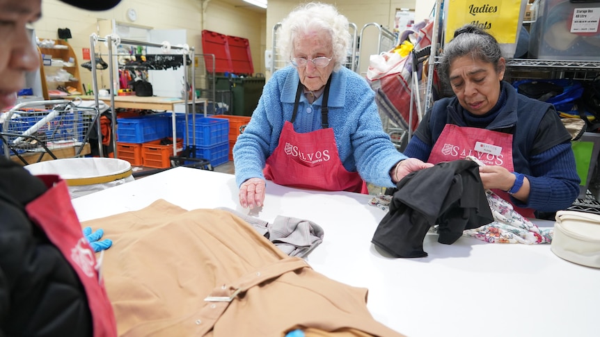 An elderly woman wearing a red Salvos' apron sorts clothes