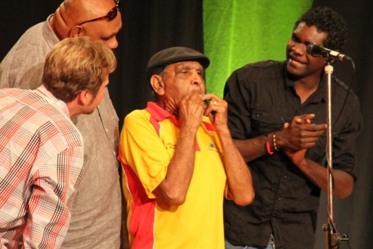 Uncle Herb plays gumleaf on stage surrounded by Indigenous musicians.