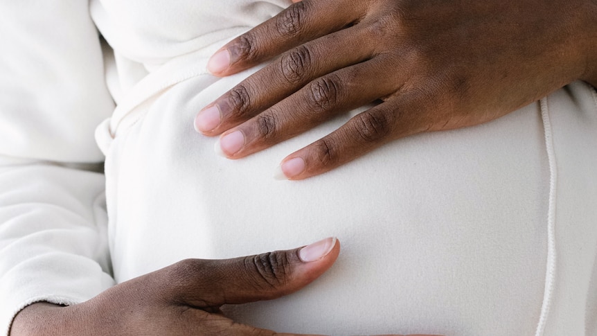 Brown hands on a white top hold a pregnant belly.