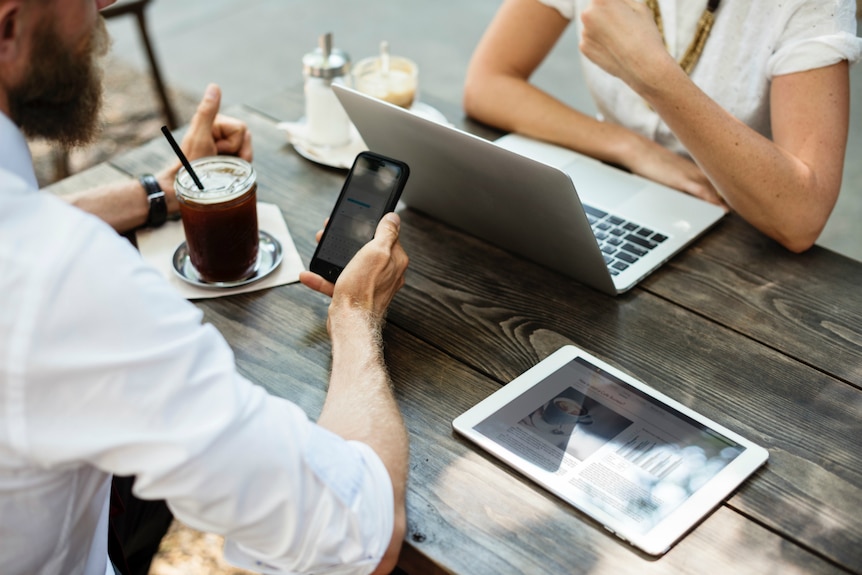 A man makes an online payment on his phone to a woman sitting opposite on her laptop.