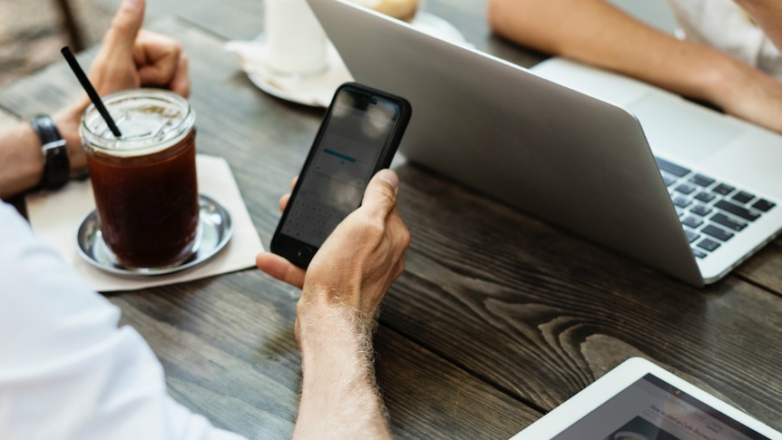 A man makes an online payment on his phone to a woman sitting opposite on her laptop.