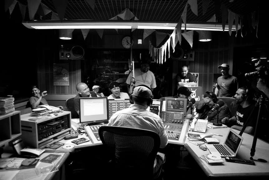A black and white photo in a radio studio, Host Hau's back is to the camera, facing him are a bunch of artists