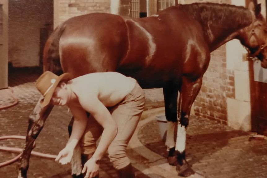 A sepia photo of a shirtless young man with a cowboy hat bending down and washing a horse