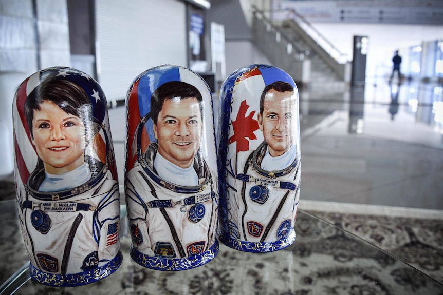 Three wooden dolls with portraits of astronauts on them sit on a table in a large room.