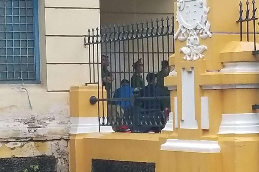People pictured from a distance through a yellow facade and bars into a court house.