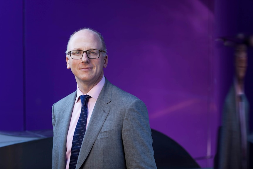 A portrait of Professor Alexander Heriot standing in front of a purple background. He's balding, wearing glasses, and a suit