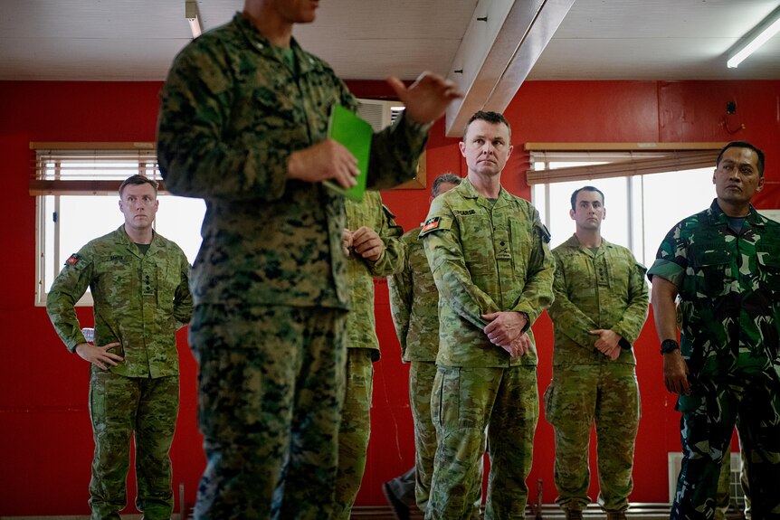 A group of men in army camouflage standing in a red room, listening to one of them talking, his hand gesturing in the air