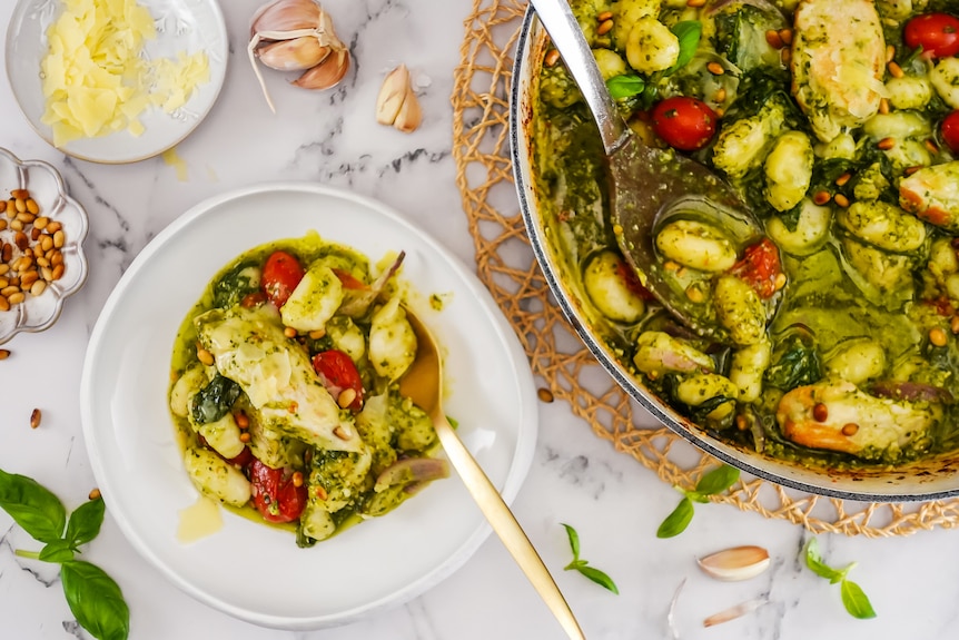 A small serving bowl of chicken pesto gnocchi with a spoon, next to a larger serving dish of it on a bench.