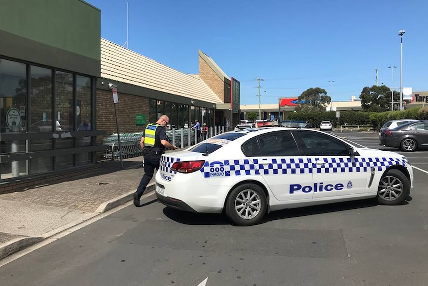 A police officer walks around the back of a police car that is parked outside a supermarket on a clear blue day.
