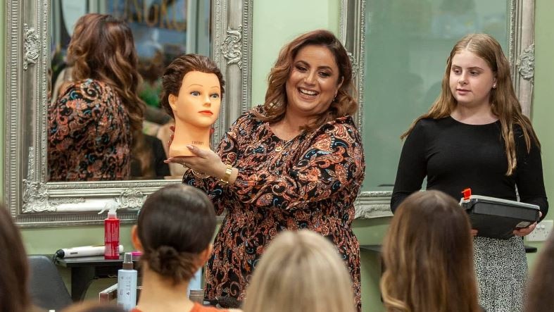 Woman in floral dress holding hairdressing manikin in front of a class with mirror in background