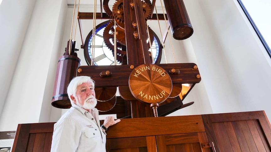 A man standing in front of a 6 metre tall wooden clock with a stain glass clock face inside a white tower.