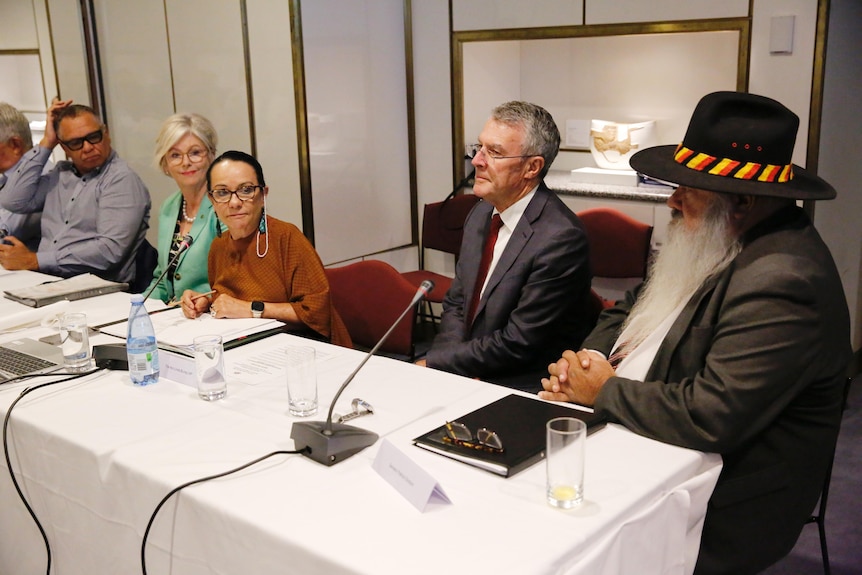 Several people sit at a table with notebooks and microphones in front of them inside parliament house.