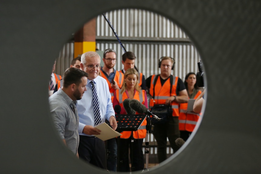 Scott Morrison is seen through the hole of a large pipe, with workers in high visibility vests standing behind him.