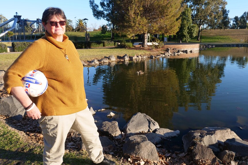 A short-haired woman stands in front of a pond with a yellow jumper and a soccer ball underneath her arm