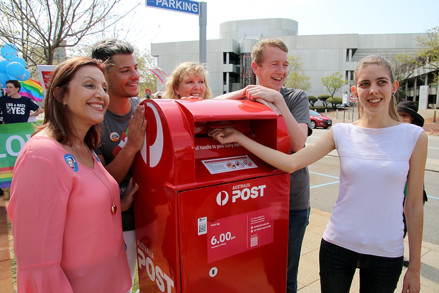 Four people gather around a post box on the side of a road while a fifth person posts a letter.