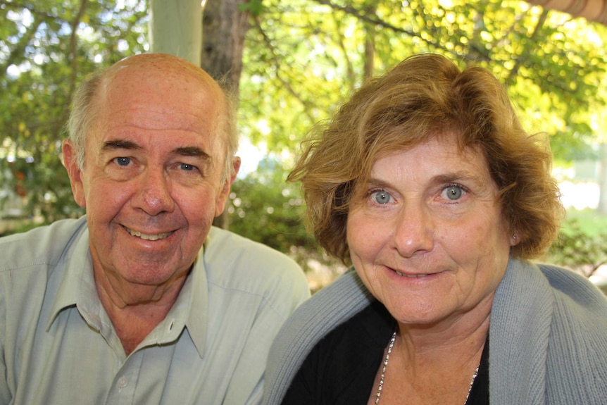 Older man and woman smile directly into camera with green trees blurred in background