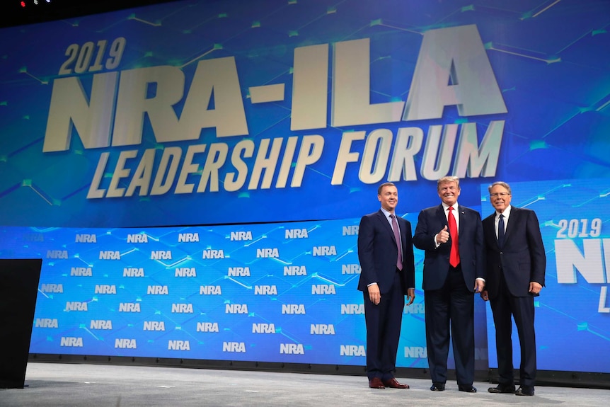 President Donald Trump stands on stage with two other men with a NRA logo behind them