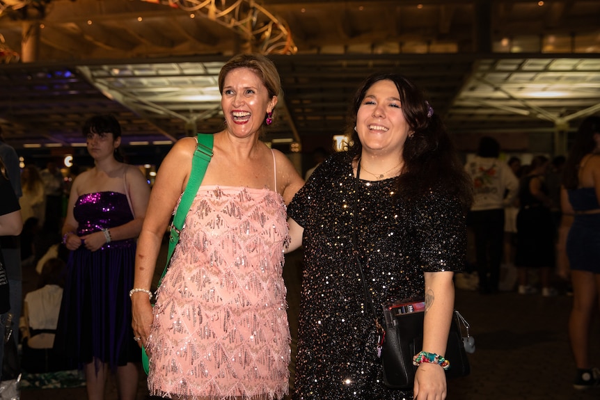 A woman in a pink sparkly dress stands with her arm around another woman wearing a sequined black dress. 