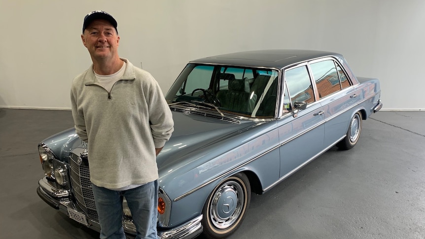A man smiles at the camera, standing in front of a vintage pale blue Mercedes-Benz car