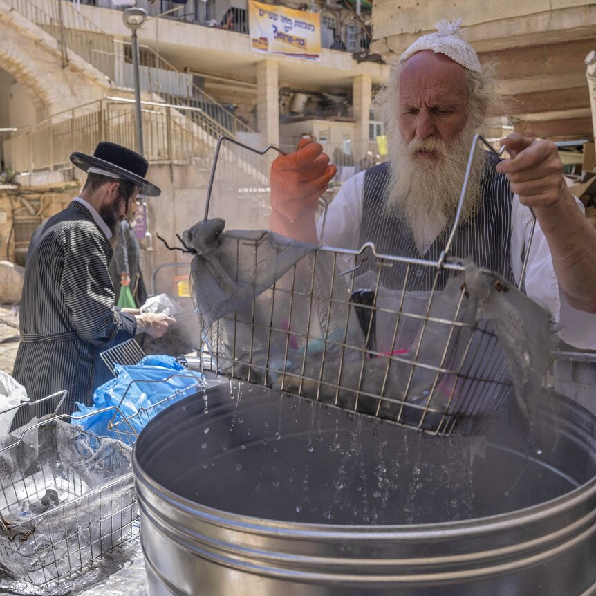 A bearded Jewish man dips cooking utensils in a large pot of boiling water.