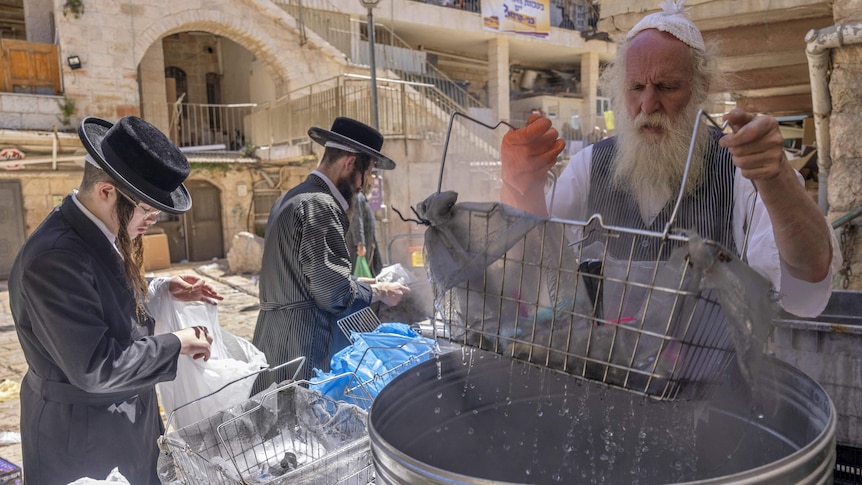 A bearded Jewish man dips cooking utensils in a large pot of boiling water.