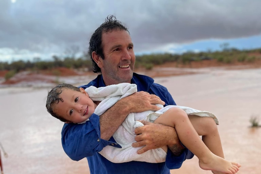 a happy man cradles his young son, who is wrapped in a towel with lots of floodwater in the background