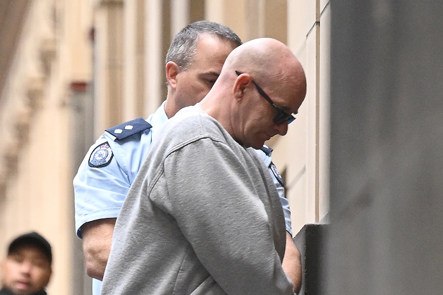 A bald man in a grey tracksuit being led by police into court.