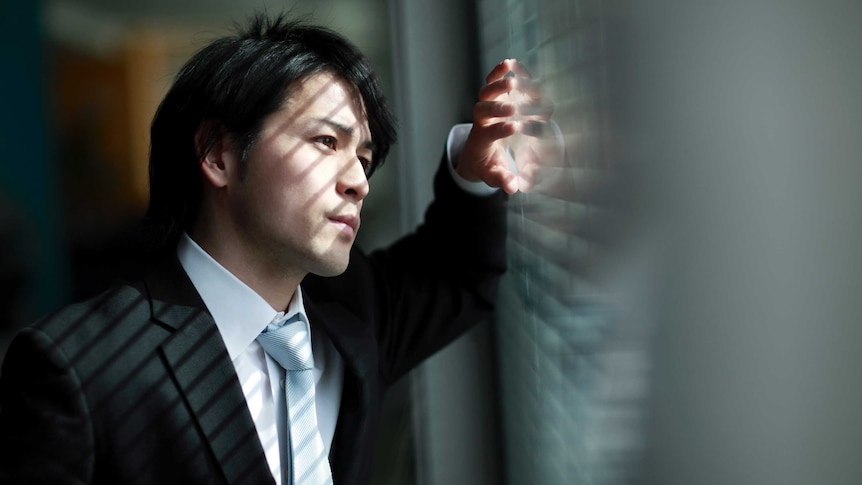 A businessman stands in his office, looking through window blinds.