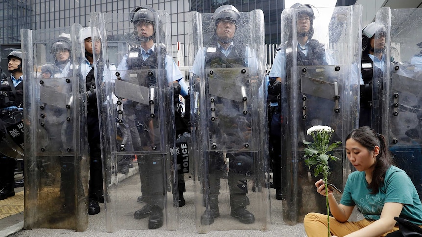 Woman in green shirt sits holding white flower in front of a line of police wearing riot gear and clear plastic shields.