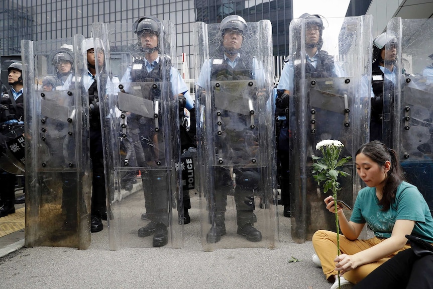 Woman in green shirt sits holding white flower in front of a line of police wearing riot gear and clear plastic shields.