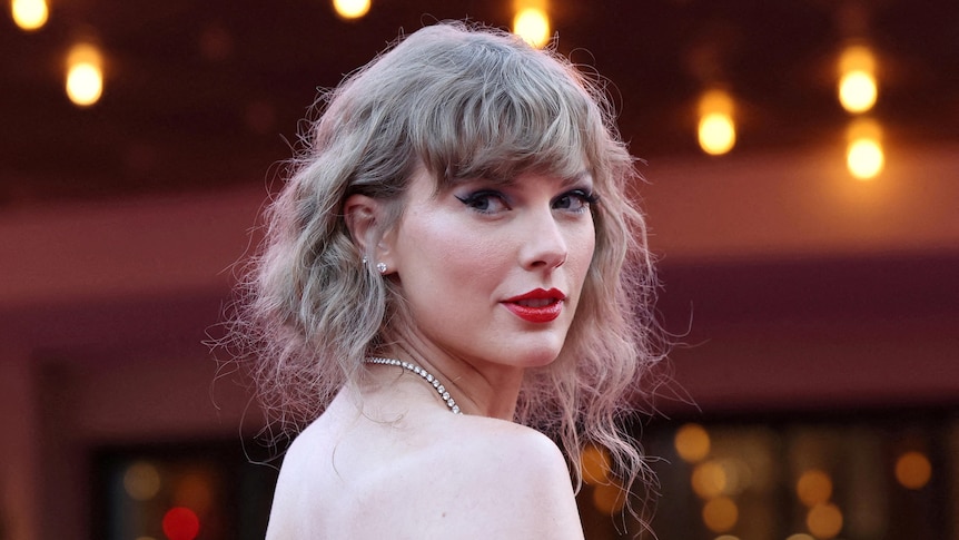 Taylor Swift looking over her bare shoulder, jewels around her neck, messy hair, red lipstick