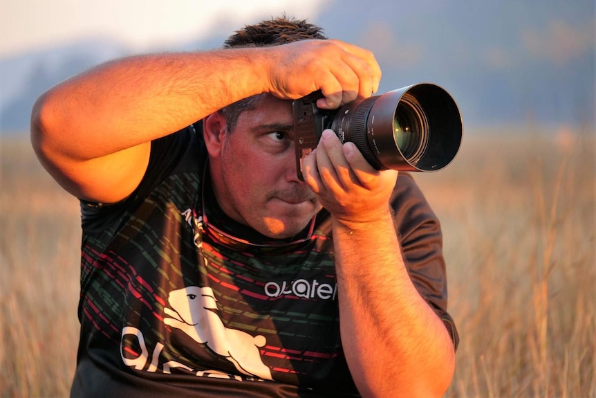 Trent White holders a camera up to his eye with a look of concentration, sun glowing on his skin, grass and mountain behind.