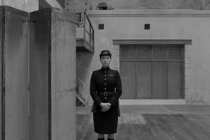 In B&W, a stern-looking policewoman in her thirties stands in a prison yard, hands carefully crossed in front of her