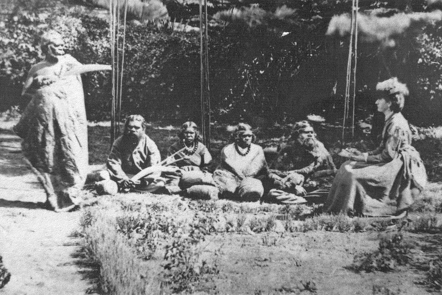 A white lady in late 1800s clothing listens to a group of indigenous people