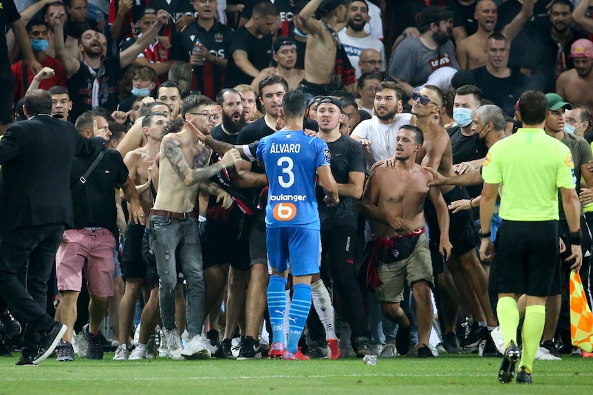 Un joueur vêtu d'un maillot de football bleu se tient devant un groupe d'individus torse nu
