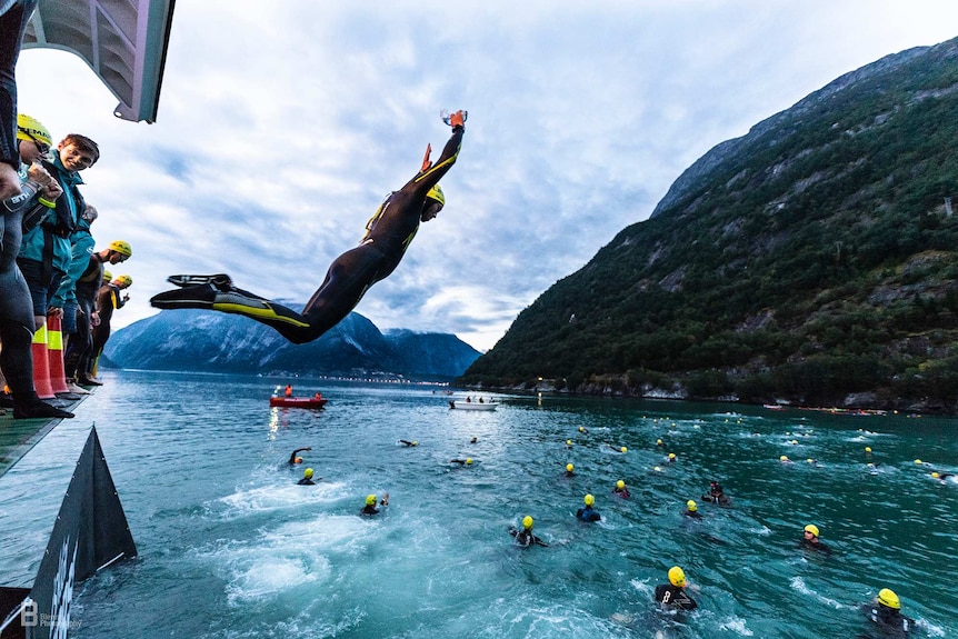 A person in a black wetsuit and yellow swimming cap jumps into a fjord surrounded by mountains.