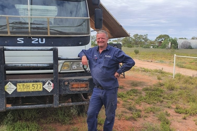 A man in blue overalls rests his elbow on a truck, a big smile on his face.