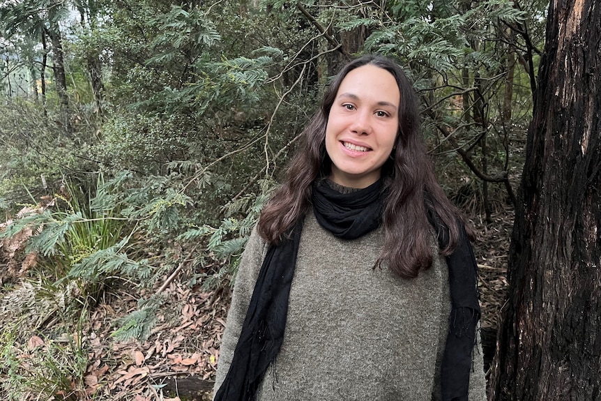 A woman with brown hair standing in a forest wearing a scarf and jumper 