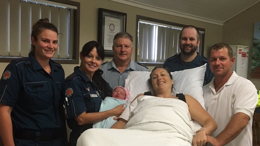 Paramedics hold newborn girl standing by mother's hospital bed.