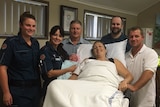 Paramedics hold newborn girl standing by mother's hospital bed.