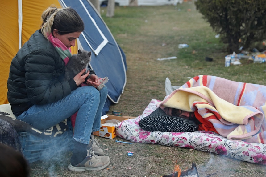 A woman holds a cat next to the site of a tent as a man sleeps on the ground.